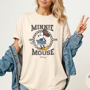 May include: Ivory-colored t-shirt featuring a vintage-style graphic of Minnie Mouse. The design includes the text "MINNIE MOUSE" and the year "1928" in a circular frame. The shirt is a comfort color and has a relaxed fit.