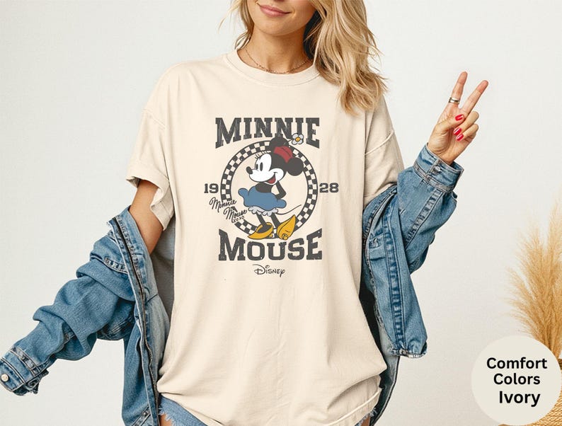 May include: Ivory-colored t-shirt featuring a vintage-style graphic of Minnie Mouse. The design includes the text "MINNIE MOUSE" and the year "1928" in a circular frame. The shirt is a comfort color and has a relaxed fit.