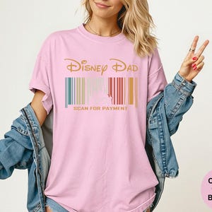 May include: Pale pink t-shirt featuring the words "Disney Dad" in gold, above a barcode design with teal, beige, red, and gold stripes. The text "SCAN FOR PAYMENT" is printed in gold below the barcode.