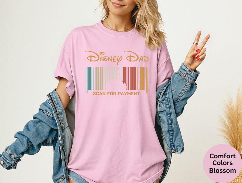 May include: Pale pink t-shirt featuring the words "Disney Dad" in gold, above a barcode design with teal, beige, red, and gold stripes. The text "SCAN FOR PAYMENT" is printed in gold below the barcode.