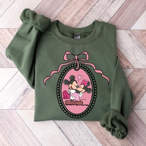 K&ouml;nnte beinhalten: Ein gr&uuml;ner Pullover mit einer rosa Schleife und einem rosa ovalen Rahmen, der Mickey Mouse und Minnie Mouse beim K&uuml;ssen zeigt. Der Rahmen ist von einem schwarzen Perlenrand umgeben.
