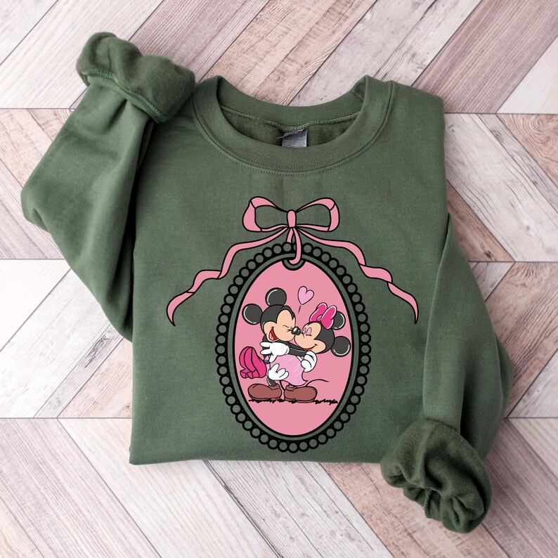 K&ouml;nnte beinhalten: Ein gr&uuml;ner Pullover mit einer rosa Schleife und einem rosa ovalen Rahmen, der Mickey Mouse und Minnie Mouse beim K&uuml;ssen zeigt. Der Rahmen ist von einem schwarzen Perlenrand umgeben.