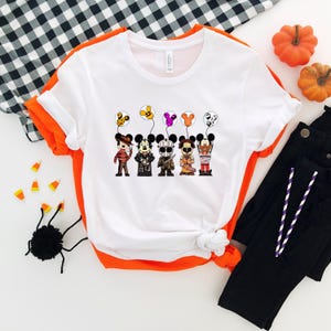 May include: White t-shirt with orange trim featuring cartoon characters dressed as Halloween villains holding balloons. The characters include Freddy Krueger, Jason Voorhees, and others. Black pants and pumpkins are in the background.