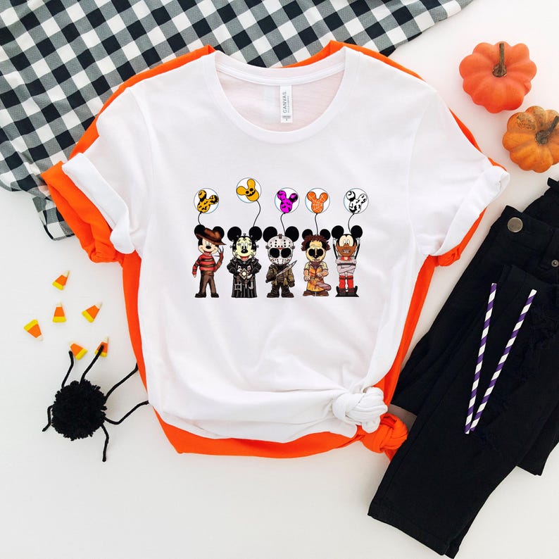 May include: White t-shirt with orange trim featuring cartoon characters dressed as Halloween villains holding balloons. The characters include Freddy Krueger, Jason Voorhees, and others. Black pants and pumpkins are in the background.