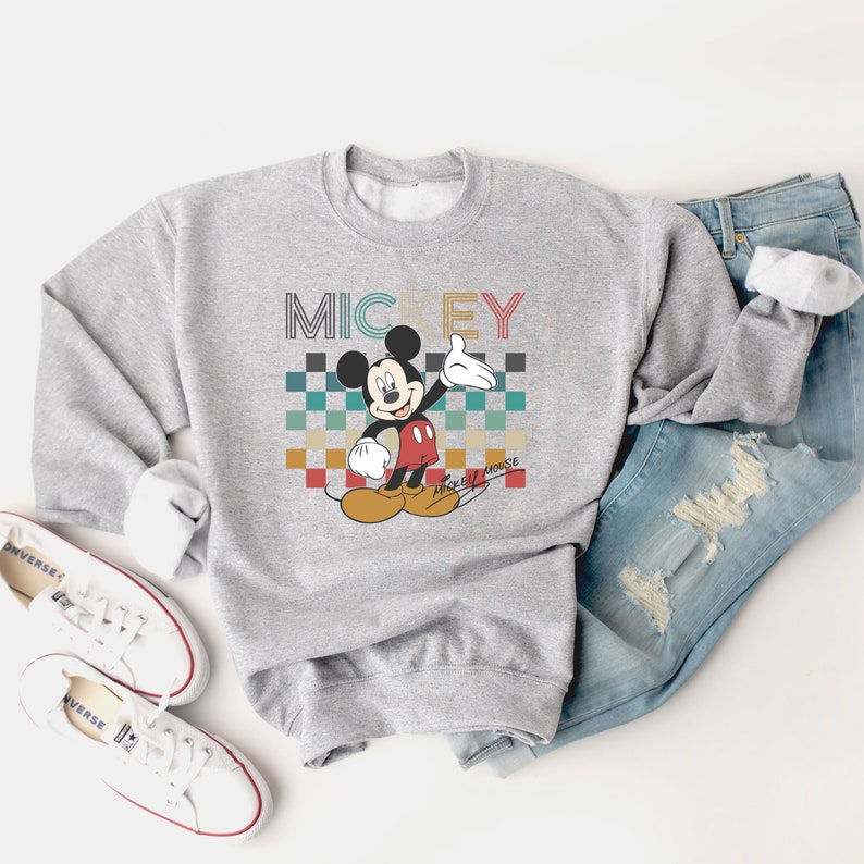 May include: A gray sweatshirt with a retro-style graphic of Mickey Mouse. The graphic features Mickey Mouse in red shorts and yellow shoes, with a checkered background and the text "MICKEY MOUSE" in a colorful font.