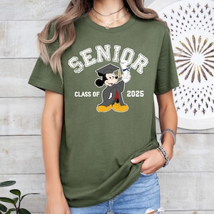 K&ouml;nnte beinhalten: Gr&uuml;nes T-Shirt mit kurzen &Auml;rmeln und einem wei&szlig;en Aufdruck von Mickey Mouse, der eine Kappe und eine Robe tr&auml;gt und ein Diplom h&auml;lt. Unter dem Aufdruck steht der Text "Senior Class of 2025".