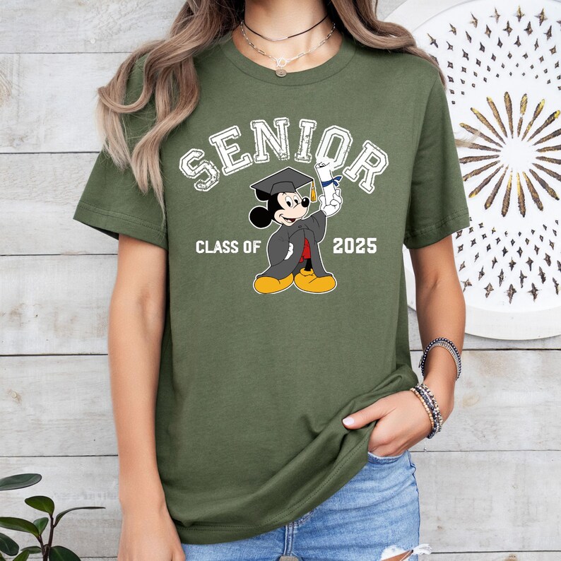 K&ouml;nnte beinhalten: Gr&uuml;nes T-Shirt mit kurzen &Auml;rmeln und einem wei&szlig;en Aufdruck von Mickey Mouse, der eine Kappe und eine Robe tr&auml;gt und ein Diplom h&auml;lt. Unter dem Aufdruck steht der Text "Senior Class of 2025".