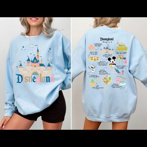 May include: Light blue sweatshirt with a graphic print of the Disneyland castle on the front and a list of things to do at Disneyland on the back. The text on the back includes "Disneyland things to do", "Churros", "Mickey's House", "Sleeping Beauty Castle", "Haunted Mansion", "It's a Small World", "Jungle Cruise", "Pirates of the Caribbean", "Space Mountain", "Frozen Ever After", "Peter Pan's Flight", "Mad Tea Party", "The Many Adventures of Winnie the Pooh", "Coco", "enjoy the magic", "and more!"