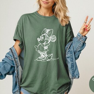 May include: Moss green t-shirt featuring a white sketch of Minnie Mouse. The shirt has short sleeves and a relaxed fit. The text "Comfort Colors Moss" is in a green circle in the bottom right corner. A denim jacket is worn over the shirt.