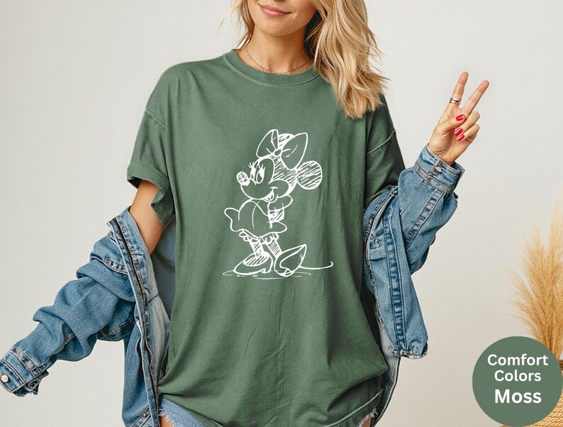 May include: Moss green t-shirt featuring a white sketch of Minnie Mouse. The shirt has short sleeves and a relaxed fit. The text "Comfort Colors Moss" is in a green circle in the bottom right corner. A denim jacket is worn over the shirt.