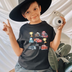 May include: A black t-shirt with a graphic print of cartoon cars and trophies. The shirt features a red, blue, and yellow color scheme.