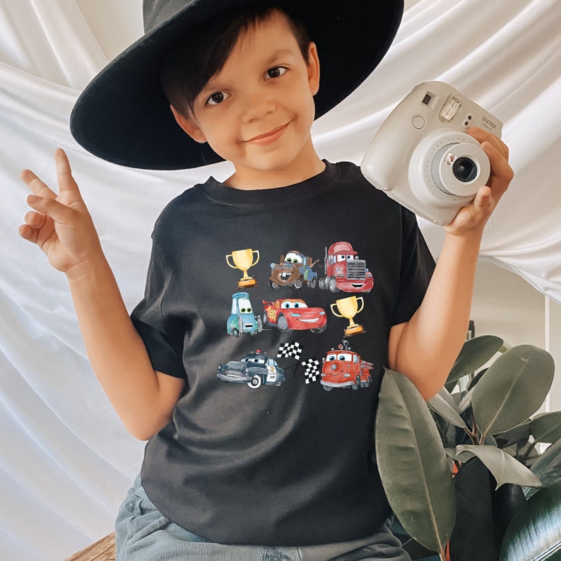May include: A black t-shirt with a graphic print of cartoon cars and trophies. The shirt features a red, blue, and yellow color scheme.