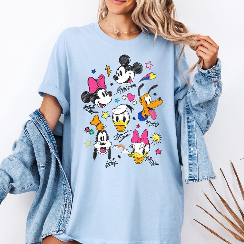 May include: Light blue t-shirt with a graphic print of Mickey Mouse, Minnie Mouse, Donald Duck, Goofy, Pluto and Daisy Duck. The characters are drawn in a cartoon style with colorful accents. The text "Mickey Mouse", "Pluto", "Goofy", and "Daisy Duck" is written beneath each character.