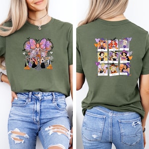 May include: Olive green t-shirt featuring a Halloween-themed graphic on the front with a bow, castle, and witch characters. The back of the shirt displays a grid of witch portraits. The shirt is paired with blue jeans.