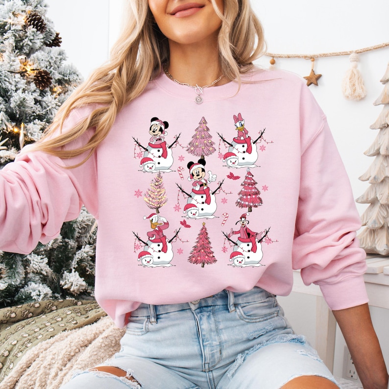 May include: A pale pink sweatshirt with a repeating pattern of snowmen featuring Disney characters such as Mickey and Minnie Mouse, Donald and Daisy Duck. The snowmen wear red scarves and hats, with pink Christmas trees and candy canes.