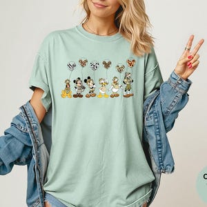 K&ouml;nnte beinhalten: Hellgr&uuml;nes Comfort Colors Bay T-Shirt mit einer Grafik von Disney-Figuren, die Luftballons mit Tiermuster halten. Die Figuren sind Pluto, Micky Maus, Minnie Maus, Donald Duck und Goofy.