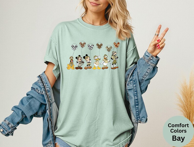 K&ouml;nnte beinhalten: Hellgr&uuml;nes Comfort Colors Bay T-Shirt mit einer Grafik von Disney-Figuren, die Luftballons mit Tiermuster halten. Die Figuren sind Pluto, Micky Maus, Minnie Maus, Donald Duck und Goofy.