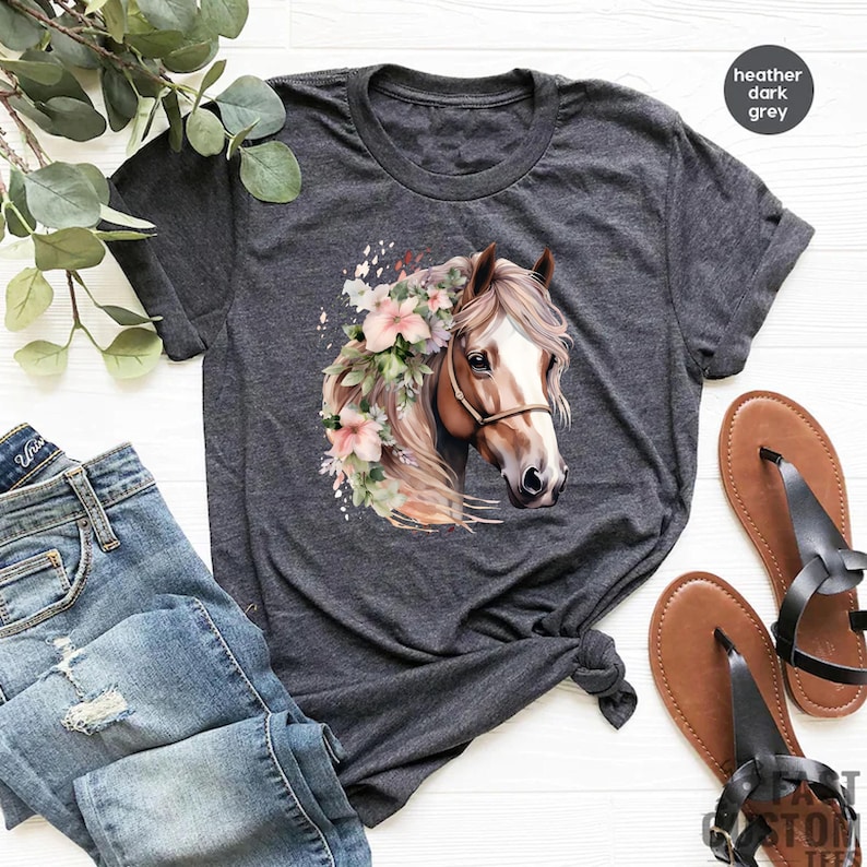 Peut inclure: T-shirt gris fonc&eacute; avec un motif de t&ecirc;te de cheval orn&eacute; d'une couronne florale. Le cheval a un visage brun et blanc et une crini&egrave;re fluide. Le t-shirt est associ&eacute; &agrave; un jean bleu et des sandales marron.