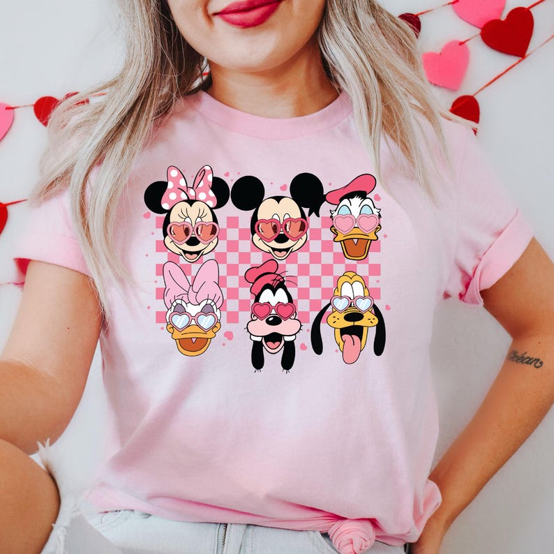 May include: Pink t-shirt with a checkered pattern featuring Mickey Mouse, Minnie Mouse, Donald Duck, Goofy, and Pluto wearing heart-shaped sunglasses.