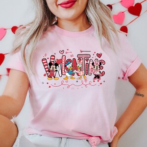 K&ouml;nnte beinhalten: Pinkes T-Shirt mit einem Valentinstags-Design mit Mickey Mouse, Minnie Mouse, Donald Duck und Daisy Duck. Der Text "Valentine" ist in Pink und Rot mit Herzen und anderen dekorativen Elementen geschrieben.
