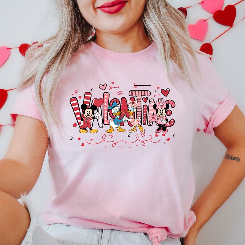 K&ouml;nnte beinhalten: Pinkes T-Shirt mit einem Valentinstags-Design mit Mickey Mouse, Minnie Mouse, Donald Duck und Daisy Duck. Der Text "Valentine" ist in Pink und Rot mit Herzen und anderen dekorativen Elementen geschrieben.