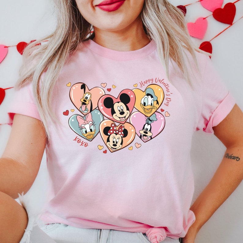 Puede incluir: Camiseta rosa con un gr&aacute;fico de Mickey Mouse, Minnie Mouse, Donald Duck, Goofy y Pluto en marcos en forma de coraz&oacute;n. El texto "Happy Valentine's Day" y "xoxo" tambi&eacute;n est&aacute;n incluidos en el dise&ntilde;o.