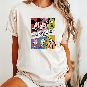 Pu&ograve; includere: Una maglietta bianca con una grafica colorata di Topolino, Minnie Mouse, Paperino, Pippo e Pluto. La grafica &egrave; in stile fumetto con ogni personaggio in una casella separata. Il testo "MICKEY & CO. EST. 1928" &egrave; sotto i personaggi.