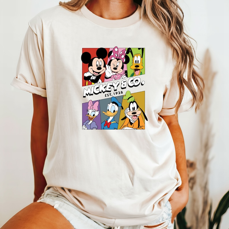 Pu&ograve; includere: Una maglietta bianca con una grafica colorata di Topolino, Minnie Mouse, Paperino, Pippo e Pluto. La grafica &egrave; in stile fumetto con ogni personaggio in una casella separata. Il testo "MICKEY & CO. EST. 1928" &egrave; sotto i personaggi.