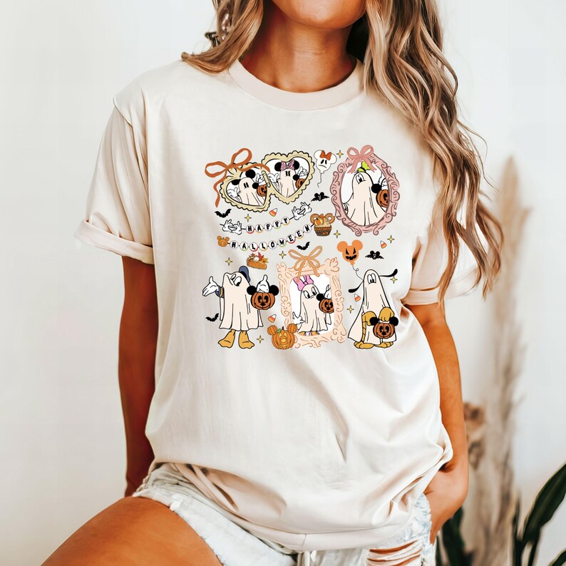 May include: Beige t-shirt featuring a Halloween-themed graphic. The design includes cartoon characters dressed as ghosts, pumpkins, and the text "HAPPY HALLOWEEN". The graphic is in shades of orange, black, and white.