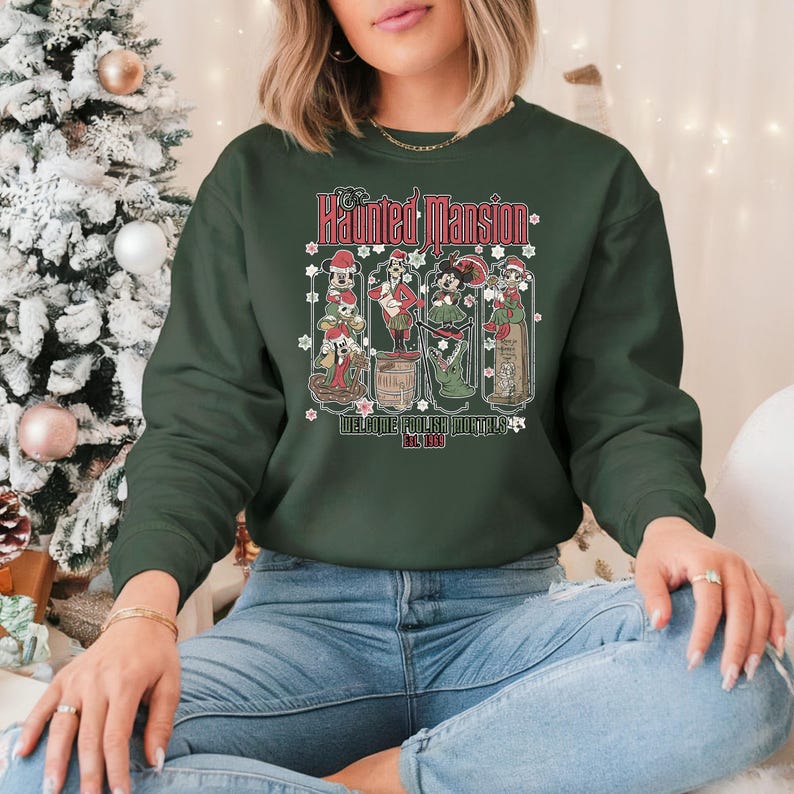 May include: A dark green sweatshirt with a festive design featuring the Haunted Mansion characters from Disney. The text on the sweatshirt reads "The Haunted Mansion Welcome Foolish Mortals Est. 1969".