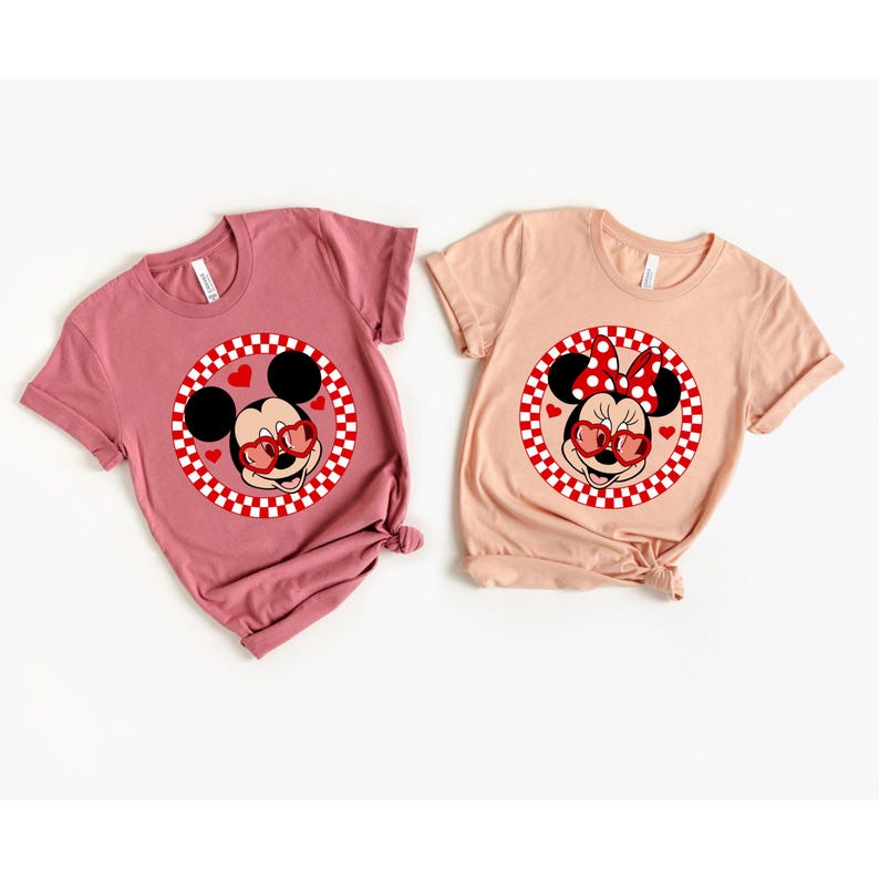 May include: Two t-shirts with a red and white checkered circle design featuring Mickey Mouse and Minnie Mouse wearing heart-shaped sunglasses. The Mickey Mouse shirt is on a pink background and the Minnie Mouse shirt is on a peach background.