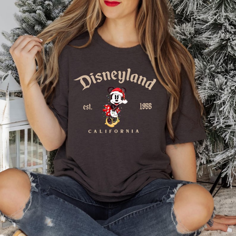 May include: A dark gray t-shirt with a vintage-style graphic of Mickey Mouse wearing a Santa hat and holding a bag of presents. The text "Disneyland est. 1955 CALIFORNIA" is printed below the graphic.