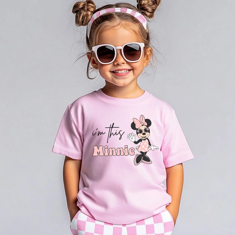 K&ouml;nnte beinhalten: Ein Kind tr&auml;gt ein rosa T-Shirt mit dem Text "I'm this Minnie" und einer Minnie-Maus-Grafik. Das Kind tr&auml;gt au&szlig;erdem eine Sonnenbrille und ein rosa-wei&szlig; gestreiftes Stirnband. Das Kind l&auml;chelt.