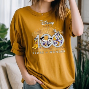 May include: A mustard yellow t-shirt with a graphic of Mickey Mouse and friends celebrating Disney's 100th anniversary. The graphic includes the text "Disney" and "100 Years of Wonder".