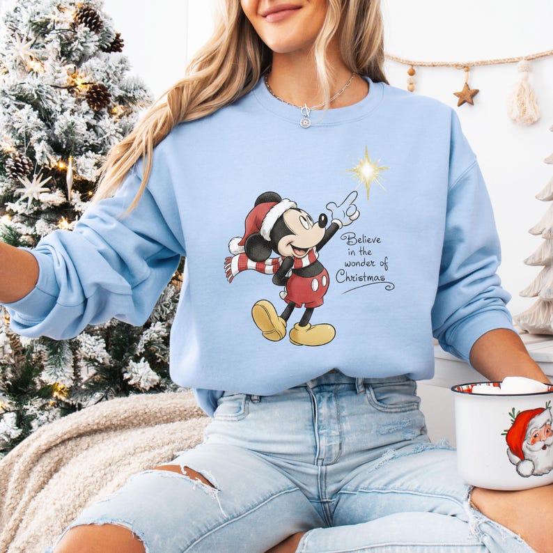 May include: Light blue sweatshirt featuring a graphic of Mickey Mouse in a Santa hat and scarf, pointing at a star. The text "Believe in the wonder of Christmas" is also on the sweatshirt. The sweatshirt is part of a Christmas-themed collection.