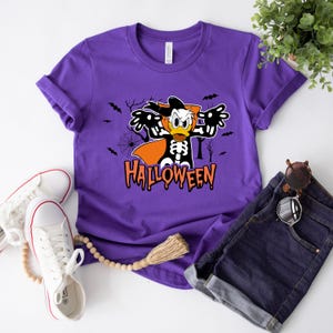 May include: A purple t-shirt featuring a Halloween-themed graphic of Donald Duck in a skeleton costume with the word "Halloween" in orange. The shirt is paired with denim shorts and white sneakers.
