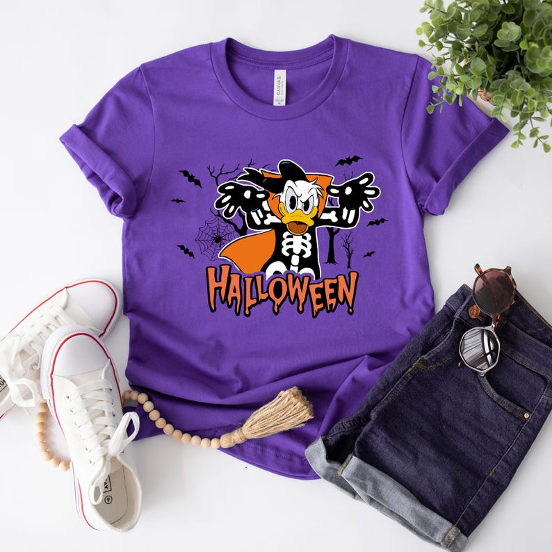 May include: A purple t-shirt featuring a Halloween-themed graphic of Donald Duck in a skeleton costume with the word "Halloween" in orange. The shirt is paired with denim shorts and white sneakers.