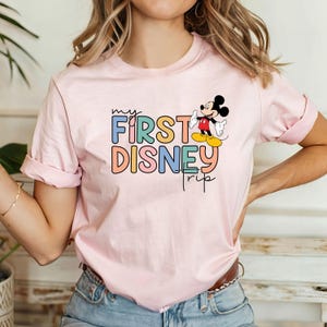 May include: Pink t-shirt with a colorful "my first disney trip" graphic and a cartoon image of Mickey Mouse.