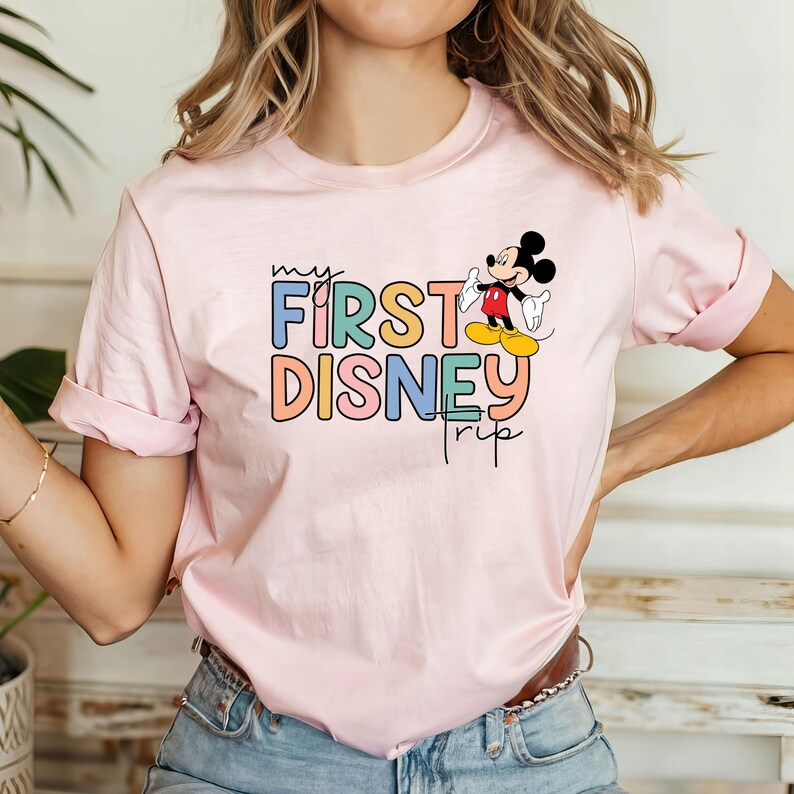 May include: Pink t-shirt with a colorful "my first disney trip" graphic and a cartoon image of Mickey Mouse.