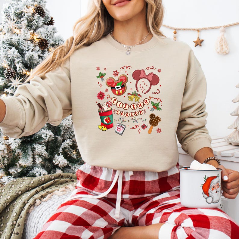 May include: Beige sweatshirt with a festive graphic featuring Mickey Mouse, Minnie Mouse, and the text "Tis the Season." The design includes Christmas elements like holly, gingerbread, and a hot cocoa cup. The person is wearing red and white plaid pajama pants.