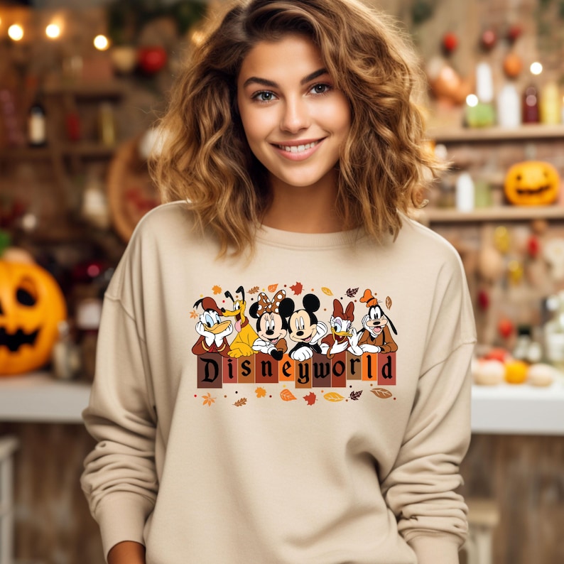 May include: Beige sweatshirt featuring a Disney World graphic with cartoon characters like Mickey and Minnie Mouse, Donald Duck, and Goofy. The graphic includes the text "Disneyworld" in a fall-themed design with leaves.