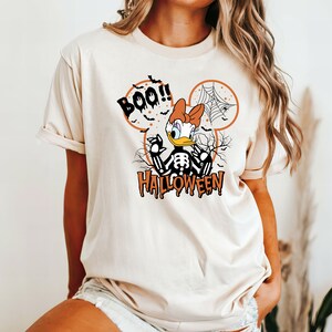 Puede incluir: Una camiseta beige con una imagen de dibujos animados de un pato esqueleto con un lazo rojo. El pato est&aacute; dentro de una silueta de oreja de Mickey con el texto "BOO!" y "HALLOWEEN" debajo.