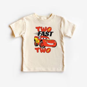 K&ouml;nnte beinhalten: Ein cremefarbenes Kleinkind-T-Shirt mit einem roten Cartoon-Rennwagen mit dem Text "TWO FAST TWO". Das Auto hat gelbe und schwarze Akzente und die Nummer 95 auf der Seite. Der Text ist in Rot und Schwarz.