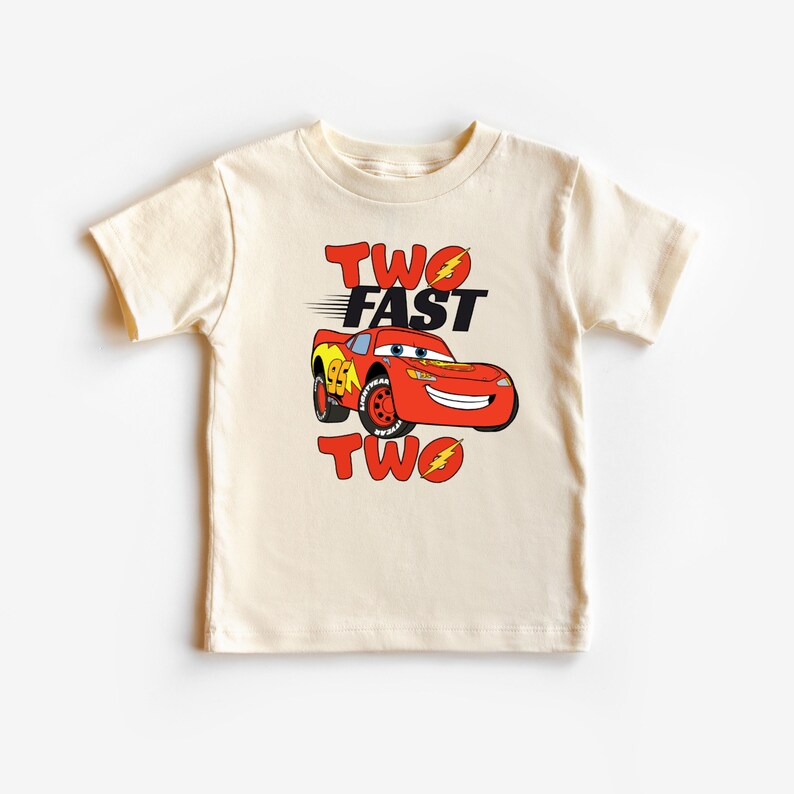 K&ouml;nnte beinhalten: Ein cremefarbenes Kleinkind-T-Shirt mit einem roten Cartoon-Rennwagen mit dem Text "TWO FAST TWO". Das Auto hat gelbe und schwarze Akzente und die Nummer 95 auf der Seite. Der Text ist in Rot und Schwarz.