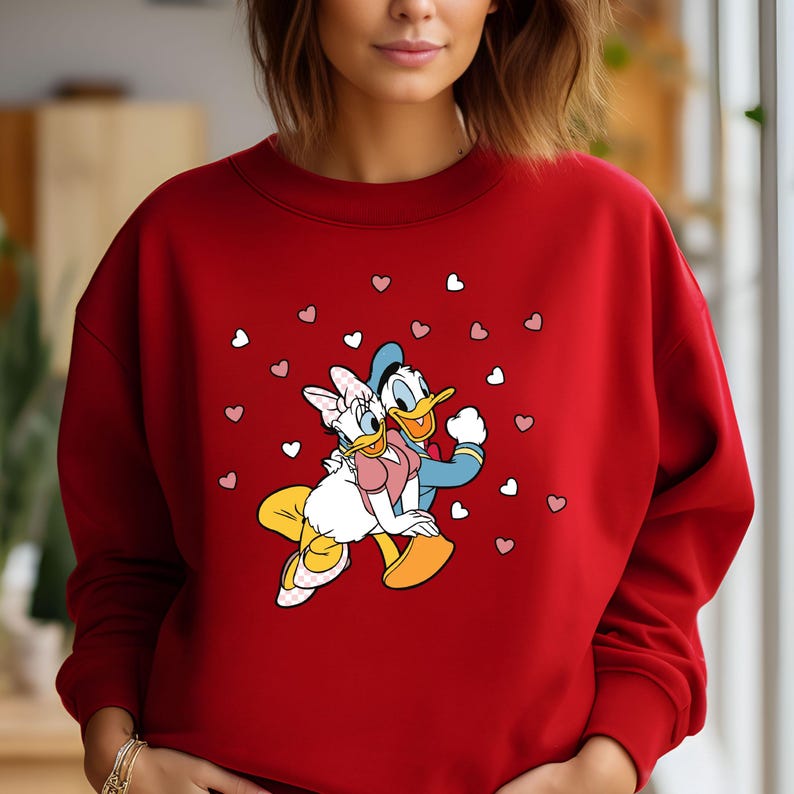 May include: Red sweatshirt with a graphic of Donald Duck and Daisy Duck holding hands surrounded by pink hearts.
