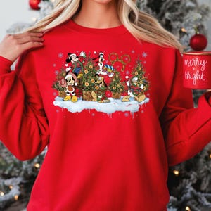 May include: Red Christmas sweatshirt featuring Mickey Mouse, Donald Duck, Goofy, and Pluto decorating Christmas trees. The characters are wearing festive attire and holding ornaments. The text "merry & bright" is visible on a mug in the background.