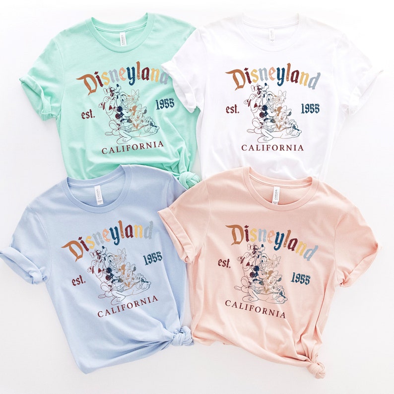 May include: Four t-shirts with a vintage Disneyland graphic featuring Mickey Mouse and friends. The shirts are in different colors: light blue, white, light green, and pink.
