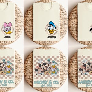 May include: Six white t-shirts with different cartoon character designs. The shirts feature Mickey Mouse, Donald Duck, Goofy, and Minnie Mouse. The shirts have a checkered pattern and the text "Mickey & Co. Est. 1928".