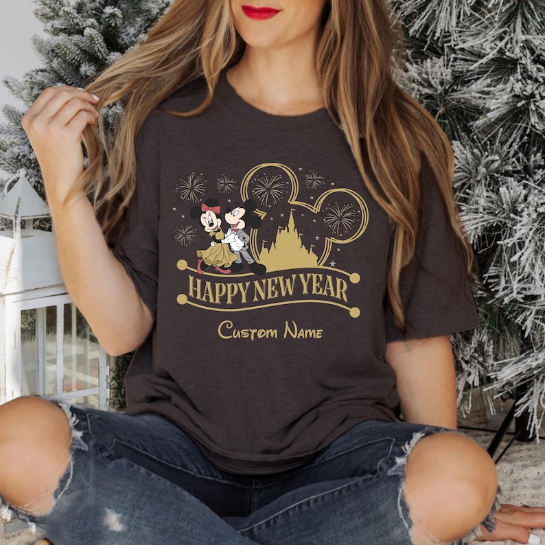 May include: A dark gray t-shirt with a gold design featuring Mickey Mouse and Minnie Mouse dancing in front of Cinderella's Castle. The design also includes fireworks and the text "Happy New Year" and "Custom Name".