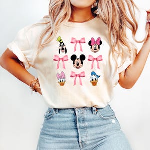 May include: A white t-shirt with a graphic print of various Disney characters, including Mickey Mouse, Minnie Mouse, Donald Duck, and Goofy. Each character is wearing a pink bow.
